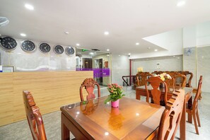 Un hall d’hôtel avec un comptoir de réception, des horloges indiquant l’heure en Chine, en Corée, au Vietnam, au Japon et aux États-Unis, un espace repas avec des chaises en bois et une table en verre, et un panneau violet indiquant que l’hôtel est situé près d’un aéroport.