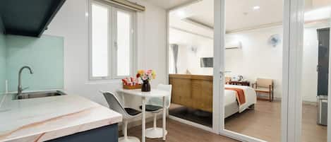 A modern hotel room with a kitchen area, a dining table with chairs, a bed with a wooden headboard, and a sliding glass door leading to a balcony.