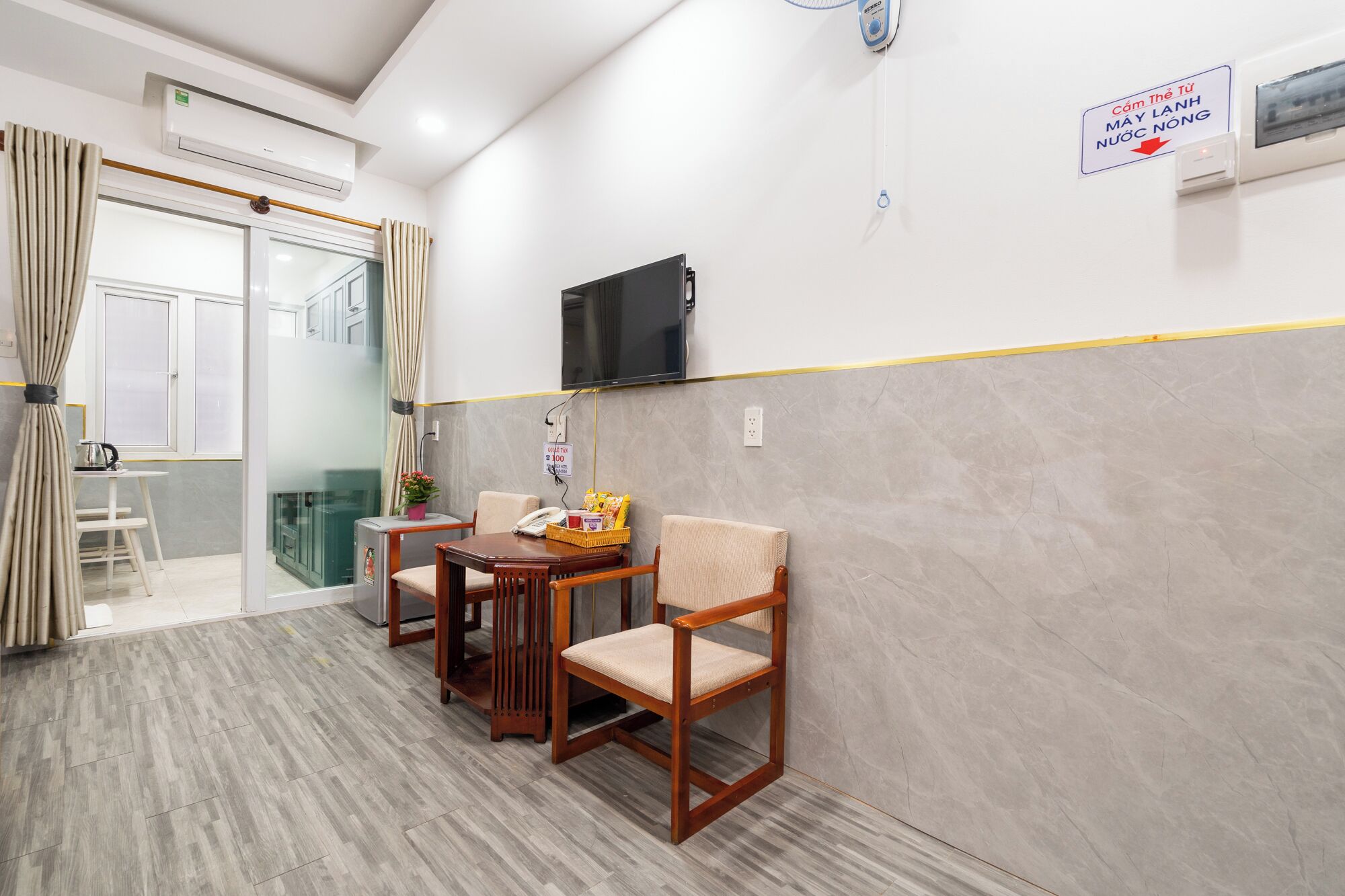 Zona de recepción de hotel con un mostrador de recepción, dos sillas de madera, una mesita con una cesta de aperitivos, un televisor montado en la pared y un cartel que indica el mostrador de registro.