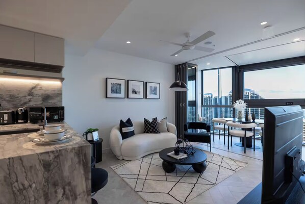 Ein modernes Wohnzimmer mit einer Marmorküche mit Kochinsel, einem schwarzen Couchtisch, einem weißen Sofa und Blick auf die Stadt durch große Fenster.