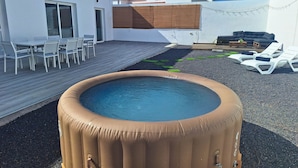 En uppblåsbar bubbelpool i en trädgård med en uteplats, utemöbler och en vit byggnad i bakgrunden.