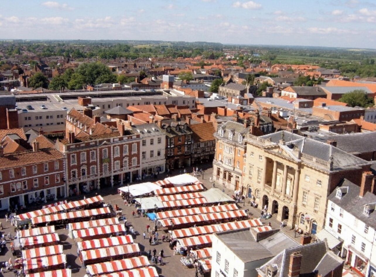 An aerial view of a historic town square with red-roofed buildings, a prominent church, and several market stalls.