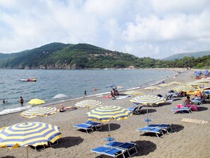 Ein Strand mit mehreren Sonnenschirmen und Liegestühlen, ein sandiger Uferstreifen und im Hintergrund Meer und Hügel.