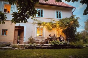 Une maison à deux étages avec un patio donnant sur le jardin, une porte en bois et un coin repas où sont assis des gens.