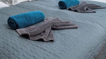 A bed with a quilted grey bedspread and two rolled towels, one blue and one brown.