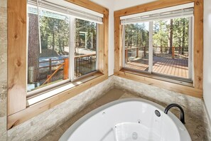 Ein Badezimmer mit einer großen Badewanne, Holzfensterrahmen und Blick auf eine Terrasse und Bäume.