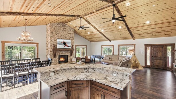A spacious living area with a stone fireplace, wooden ceiling beams, and a large window overlooking a forest. - Custom Log Style Cabin! Foosball! BBQ! Decks! Fenced Yard! (Big Bear Lake)