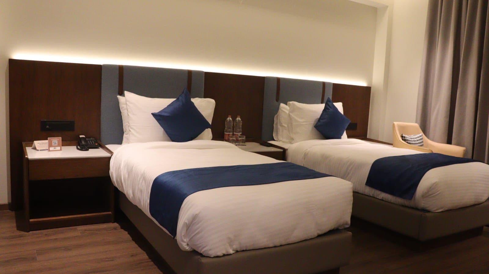 A hotel room with two beds, each with a pillow and a blue accent. There is a bedside table with a phone and a water bottle.