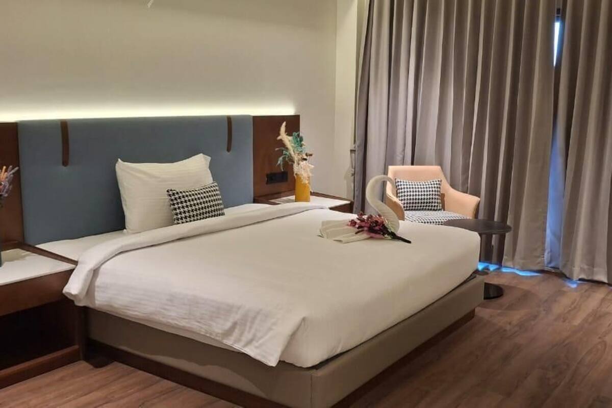 A hotel room with a large bed, a bedside table with a vase, a chair with a decorative pillow, and a view of the outside through curtains.