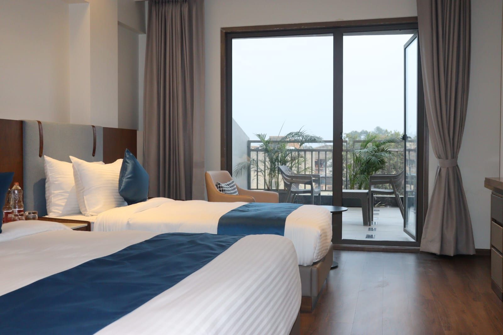 Habitación de hotel con dos camas, un balcón con mobiliario exterior y vistas a la ciudad.