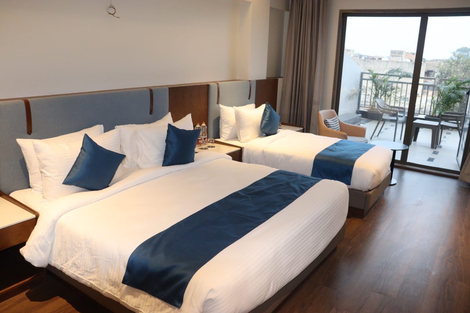 A hotel room with two beds, a balcony with outdoor seating, and a large window.