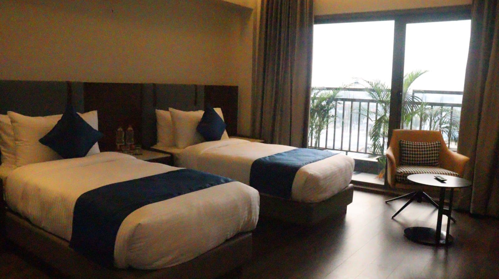 A hotel room with two beds, a small table, a chair, and a balcony with a view.