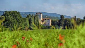 Un edificio storico in pietra con una torre, circondato da vegetazione rigogliosa e colline sullo sfondo.