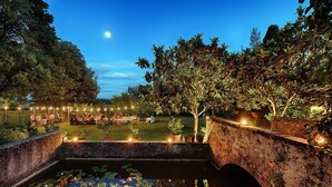 Un giardino con un ponte di pietra sopra uno stagno, circondato da alberi e luci decorative.