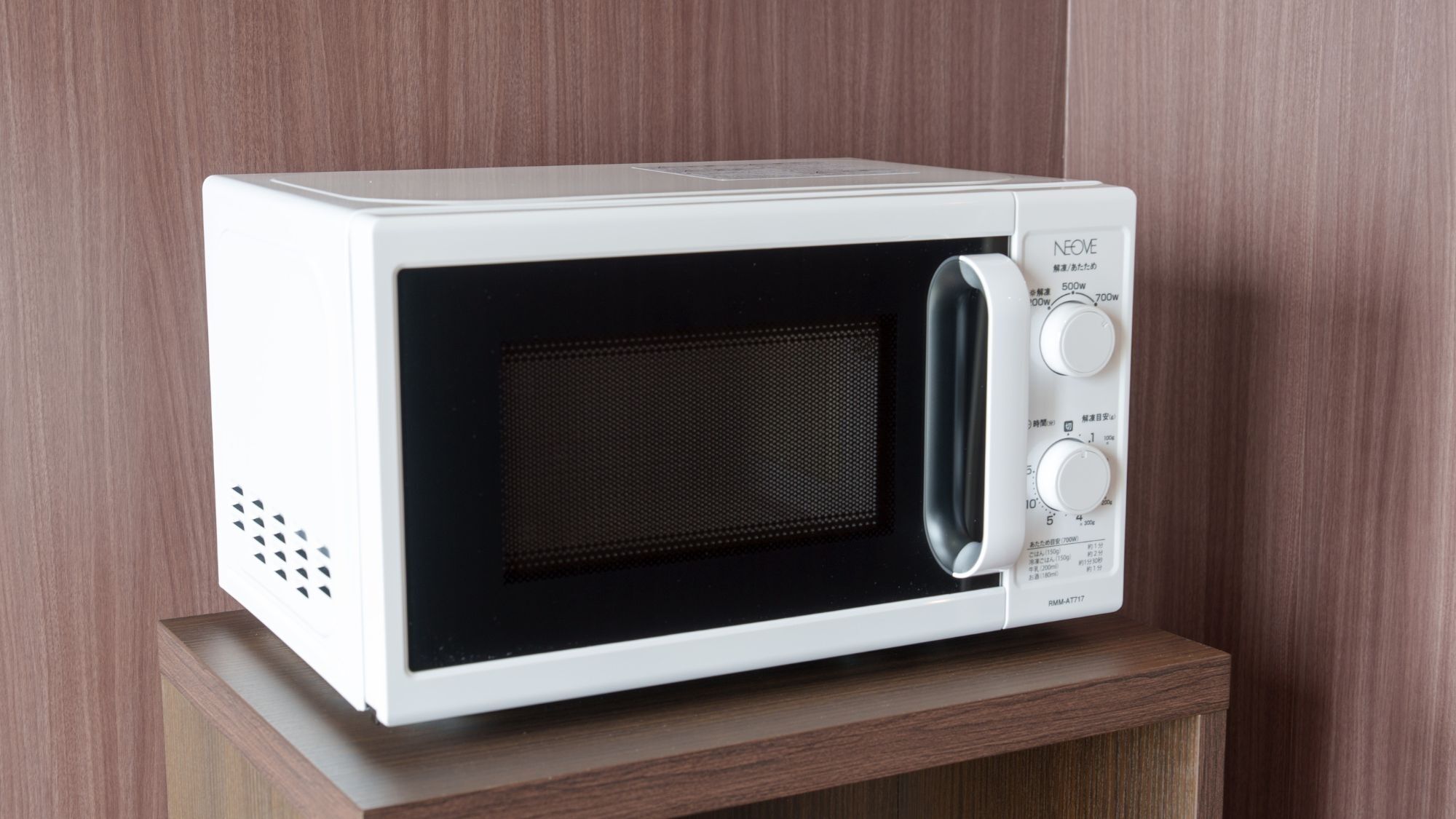 A white microwave oven with a control panel, placed on a wooden shelf against a wooden wall.