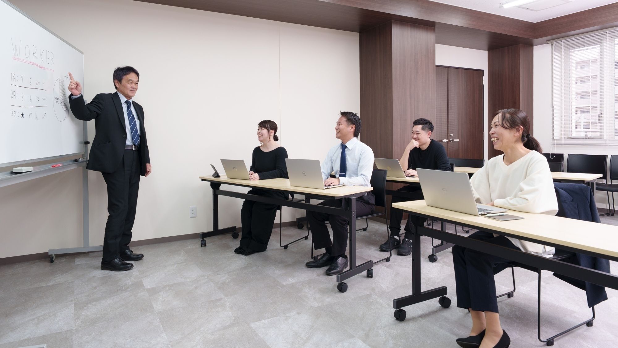 Una sala de conferencias con una pizarra, un presentador y asistentes sentados en mesas con computadoras portátiles.