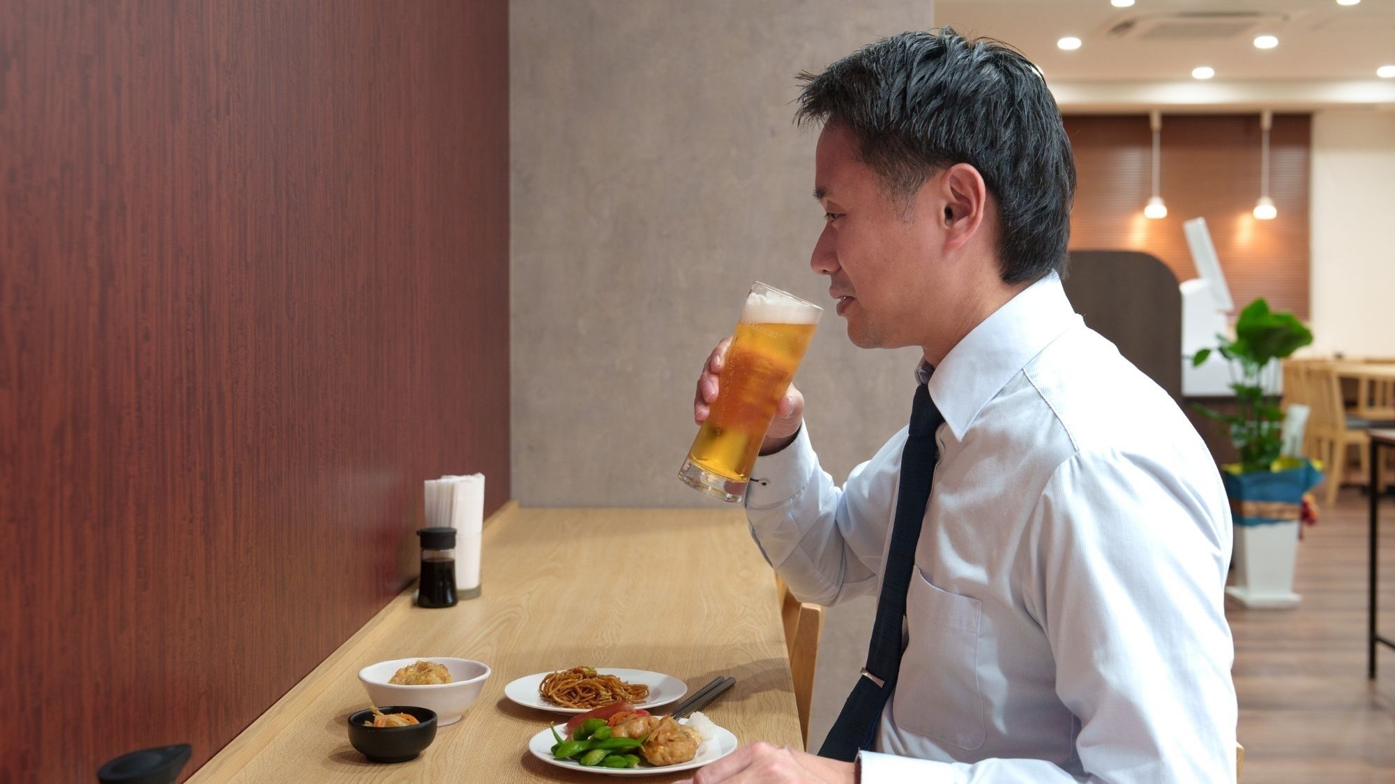 Iemand zit aan een houten tafel met een maaltijd en een glas bier, in een ruimte met een houten wand en hangende lampen.