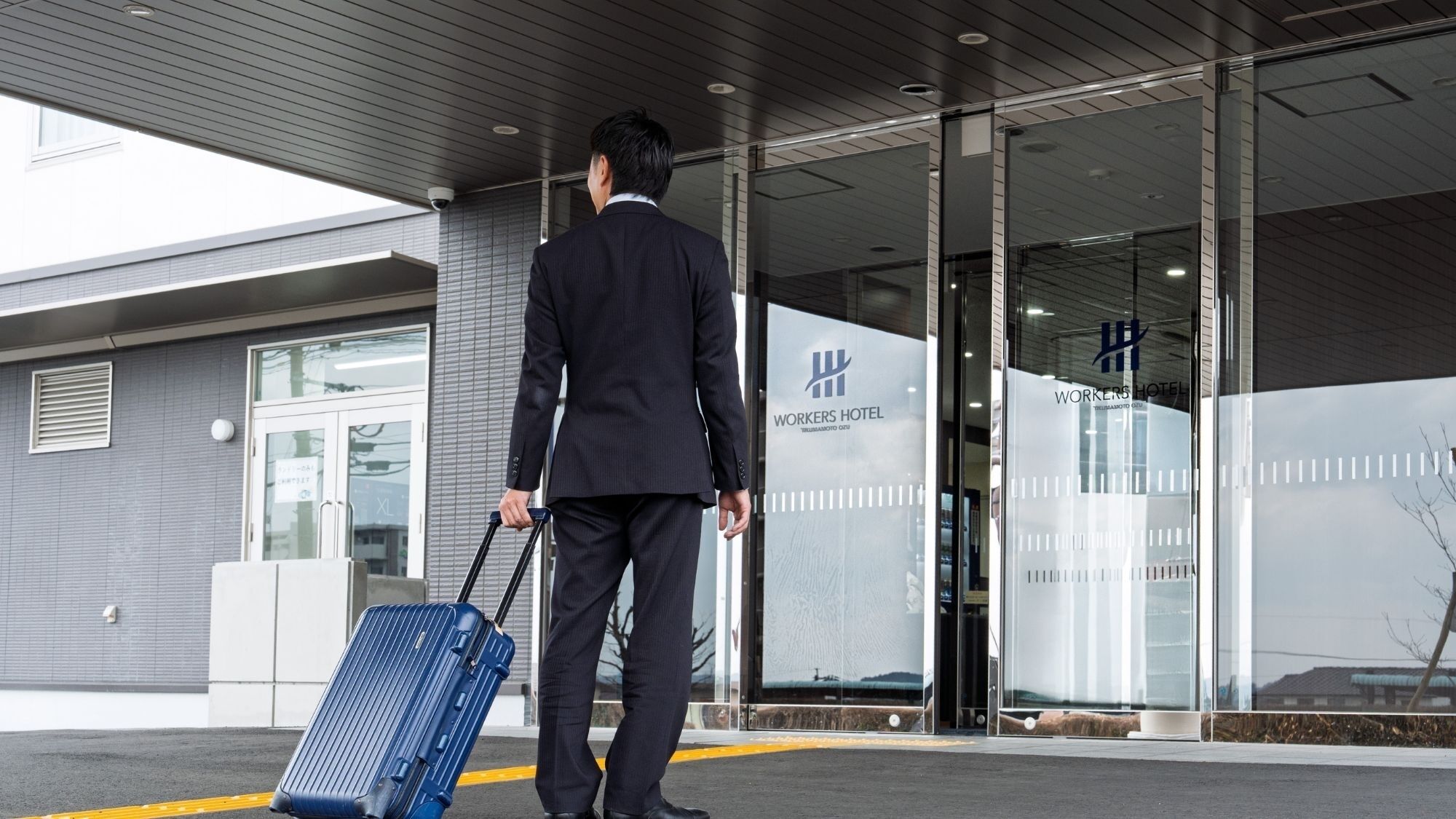 A man in a suit walking towards a hotel entrance with a blue suitcase.
