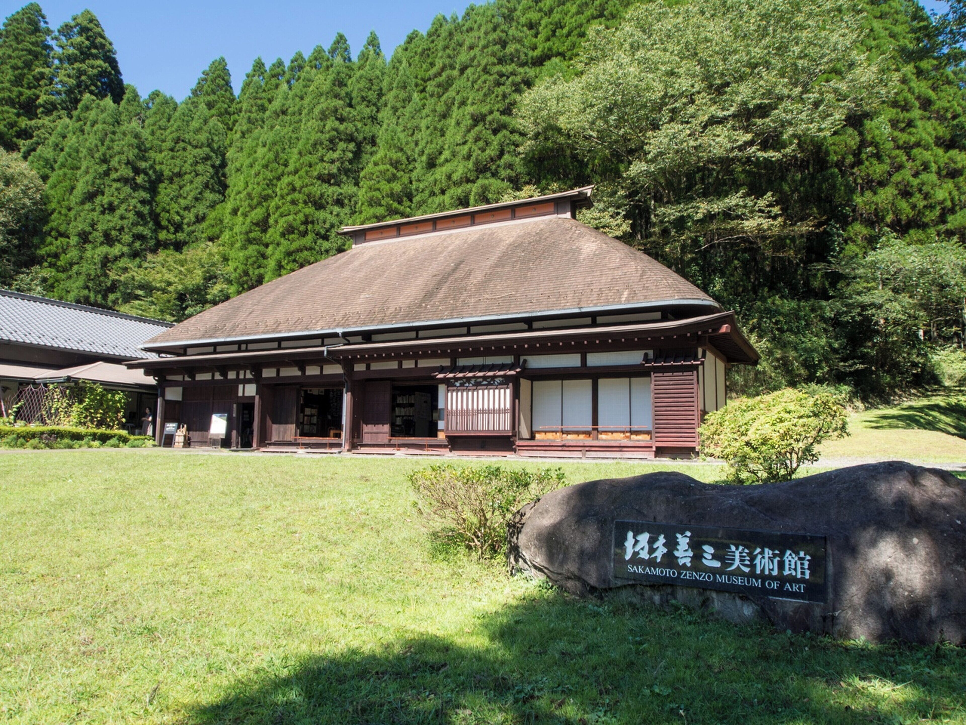 Un bâtiment japonais traditionnel, doté d’un toit en tuiles et d’une structure en bois, entouré d’une végétation luxuriante et agrémenté d’une pancarte en pierre devant l’entrée.
