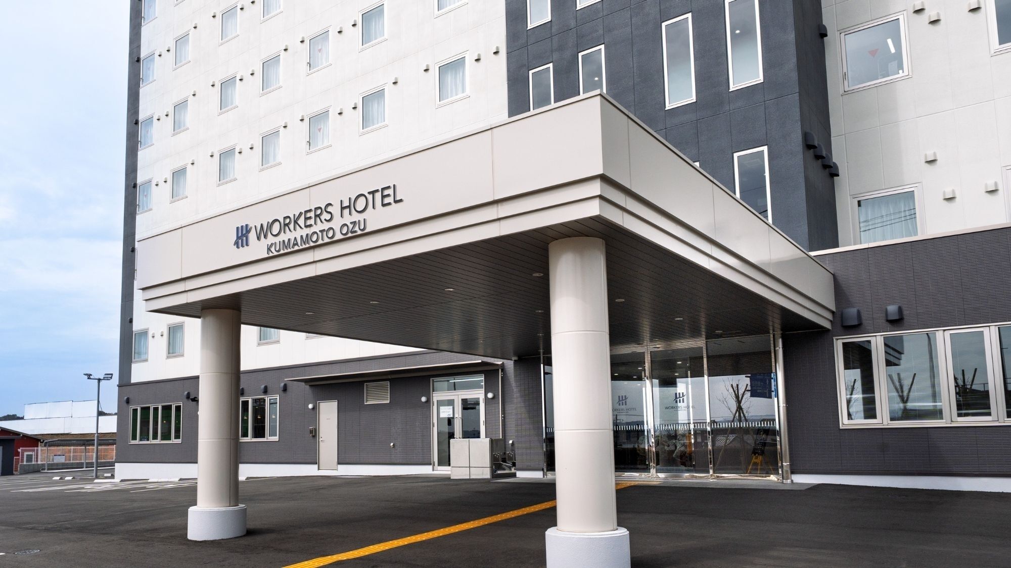 Fachada de un hotel con un corredor cubierto, amplias ventanas y un cartel que dice 'WORKERS HOTEL KUMAMOTO OZU'.