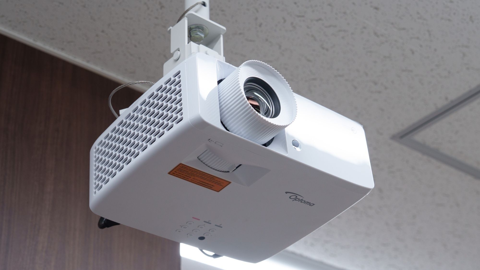 Proyector blanco montado en una pared con una estructura de techo, un cable de alimentación y una etiqueta con texto.