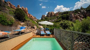 Ein Poolbereich mit Liegestühlen und einem Sonnenschirm, umgeben von felsigem Gelände und Grünflächen.