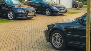 Een meerlaags woongebouw met een bakstenen gevel, een hekwerk en twee geparkeerde auto's op de oprit.