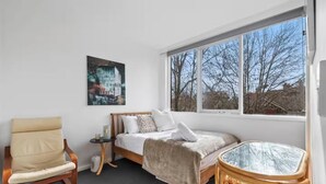 Ein Schlafzimmer mit einem Bett, einem Sessel, einem kleinen Tisch und einem Fenster mit Blick auf Bäume.