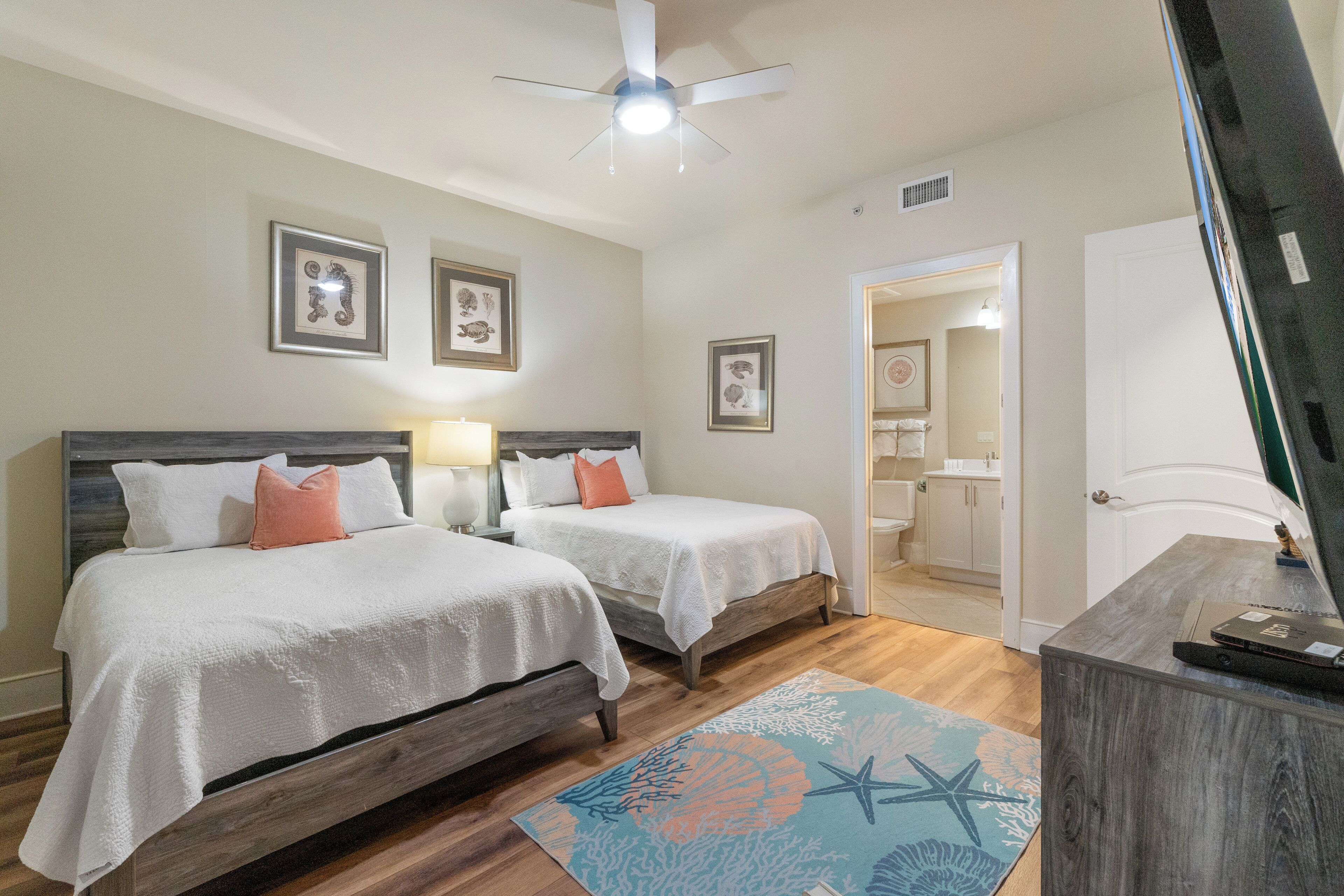 A bedroom with two beds, a ceiling fan, framed pictures on the wall, and a bathroom visible through an open door.