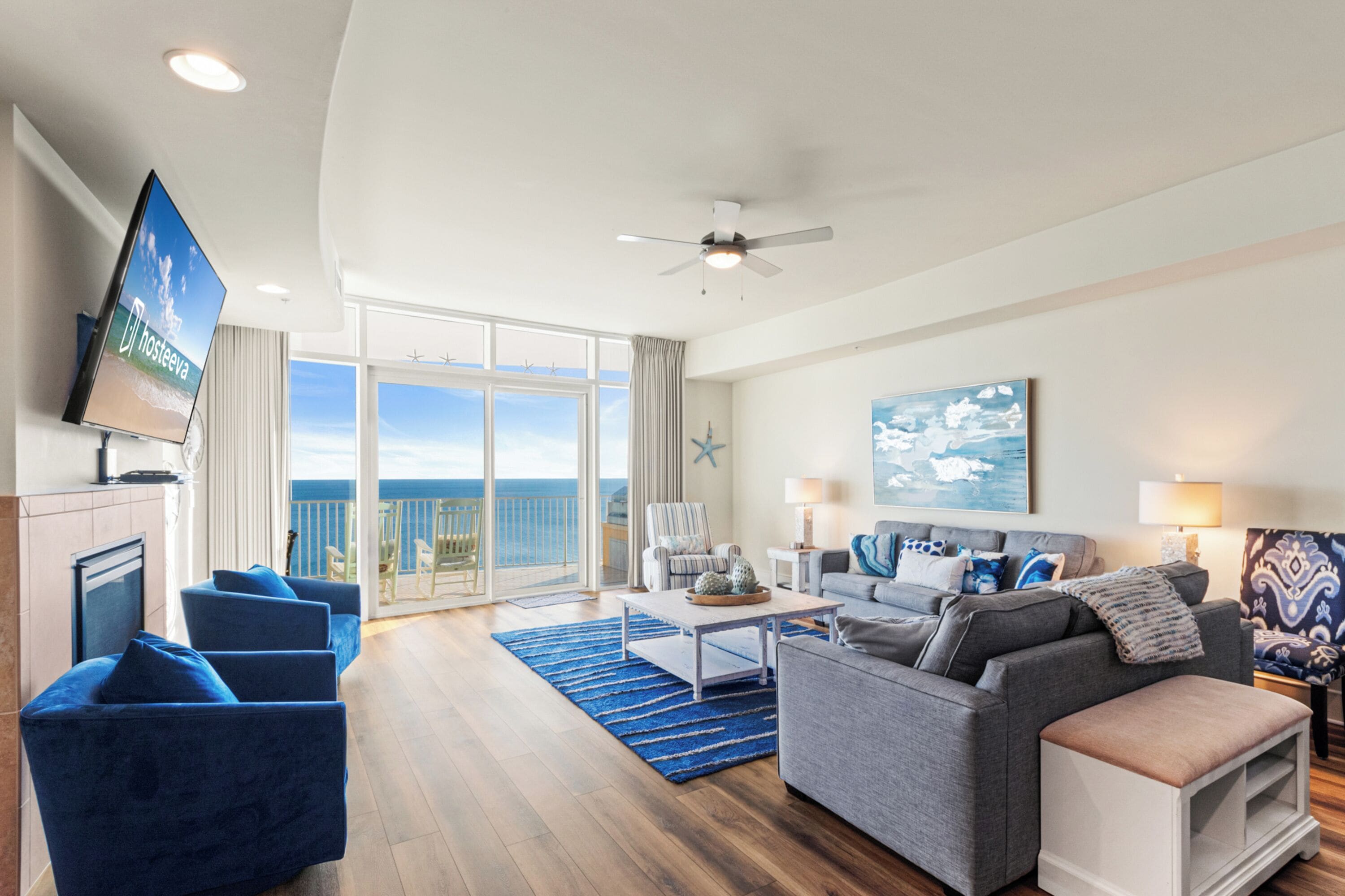 A spacious living room with a large TV, a ceiling fan, a sofa, a coffee table, and a view of the ocean through the balcony doors.