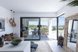 A modern living room with a sofa, a dining table, and a large window offering a view of the outdoors. - [NEW]Villa Es-Hi w Private Pool & Scenic Sea View (Kalyves)