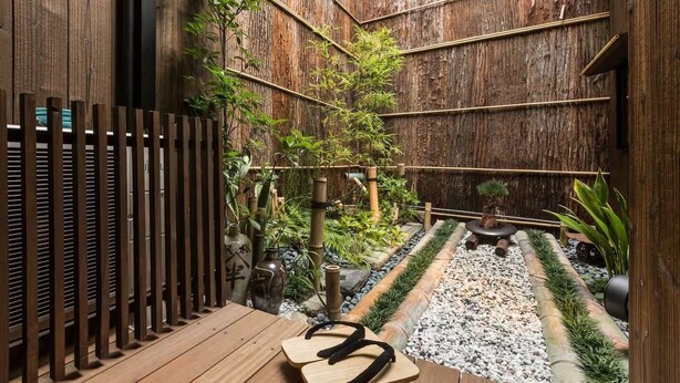 Een houten terras met een kleine tafel en stoelen, omringd door een bamboomschutting en diverse planten.