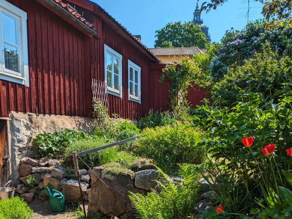 Una casa di legno rossa con un giardino antistante, un muro in pietra e una chiesa sullo sfondo.