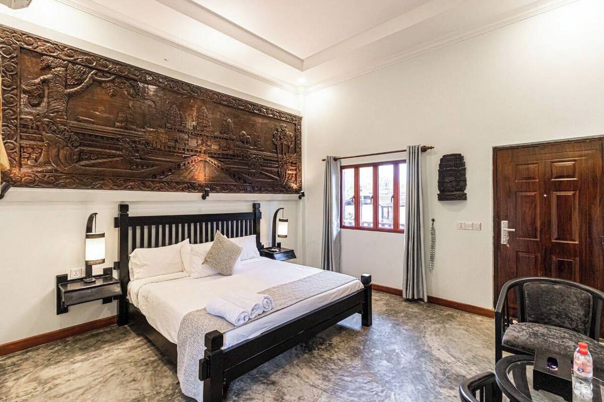 A neatly arranged hotel room with a large bed, a wooden headboard, a wall-mounted artwork, a window with curtains, and a small table with a chair.