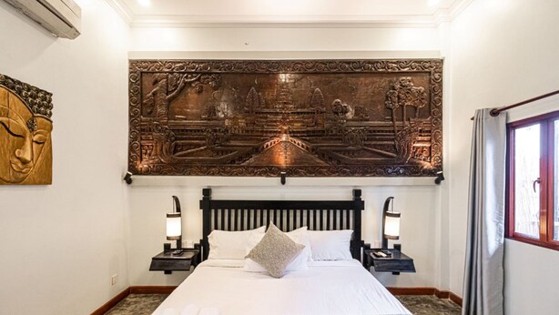 A hotel room with a large bed, two bedside tables with lamps, a wall-mounted artwork depicting a temple scene, and a window with curtains. - Teko Island (Siem Reap)
