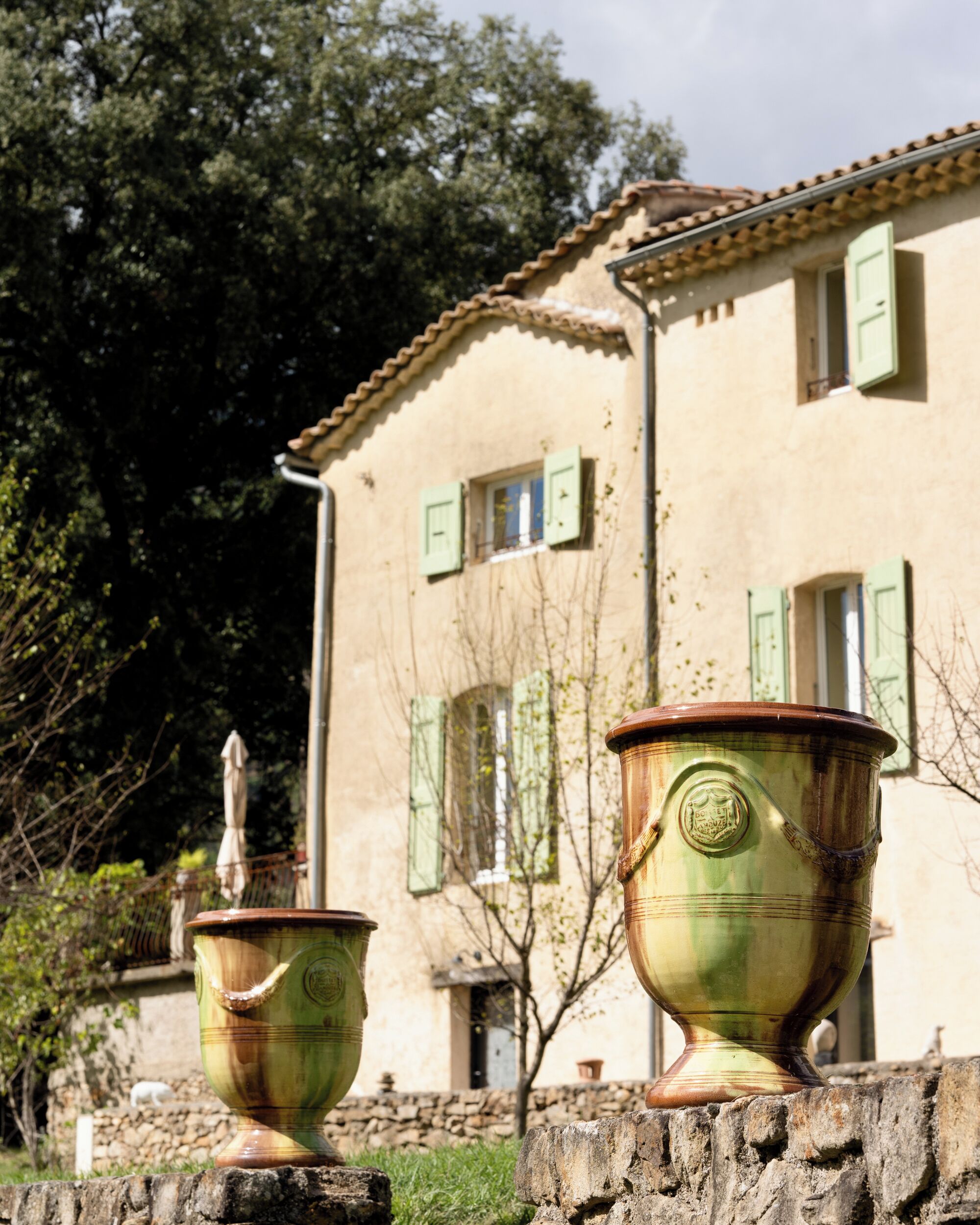 Two large ceramic pots in front of a building with green shutters and a stone wall.