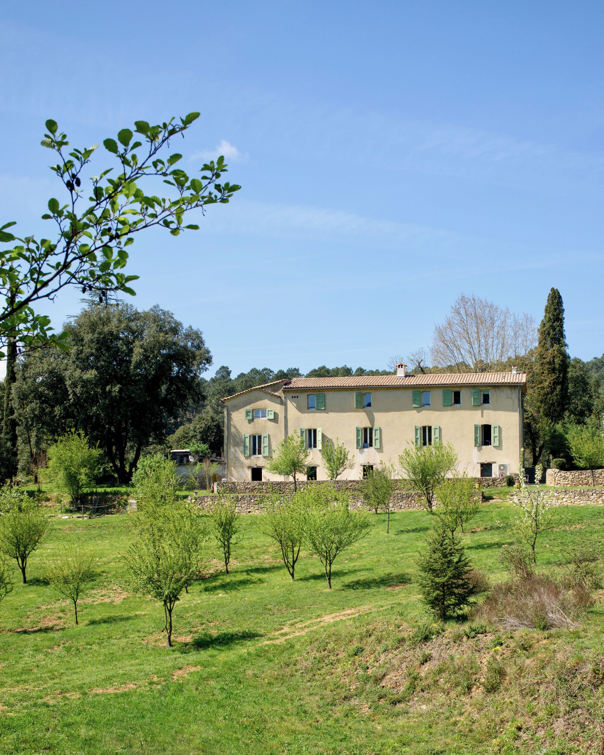A two-story building with a stone facade, multiple windows with shutters, and a surrounding grassy area with young trees.