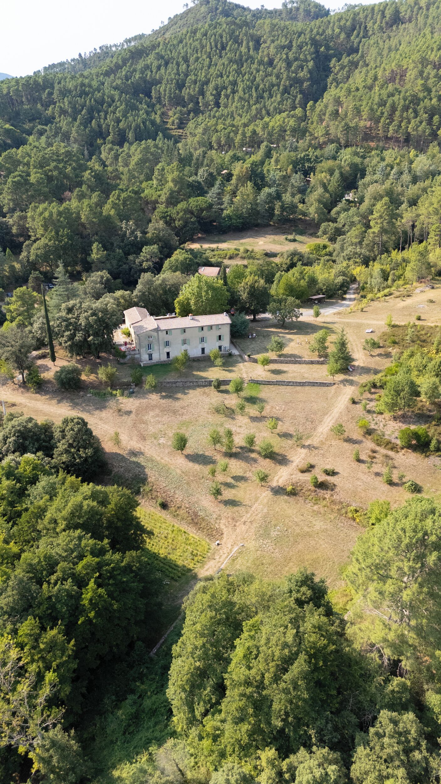 A large, two-story building with a flat roof, surrounded by a mix of dense and sparse vegetation, set against a backdrop of hills.