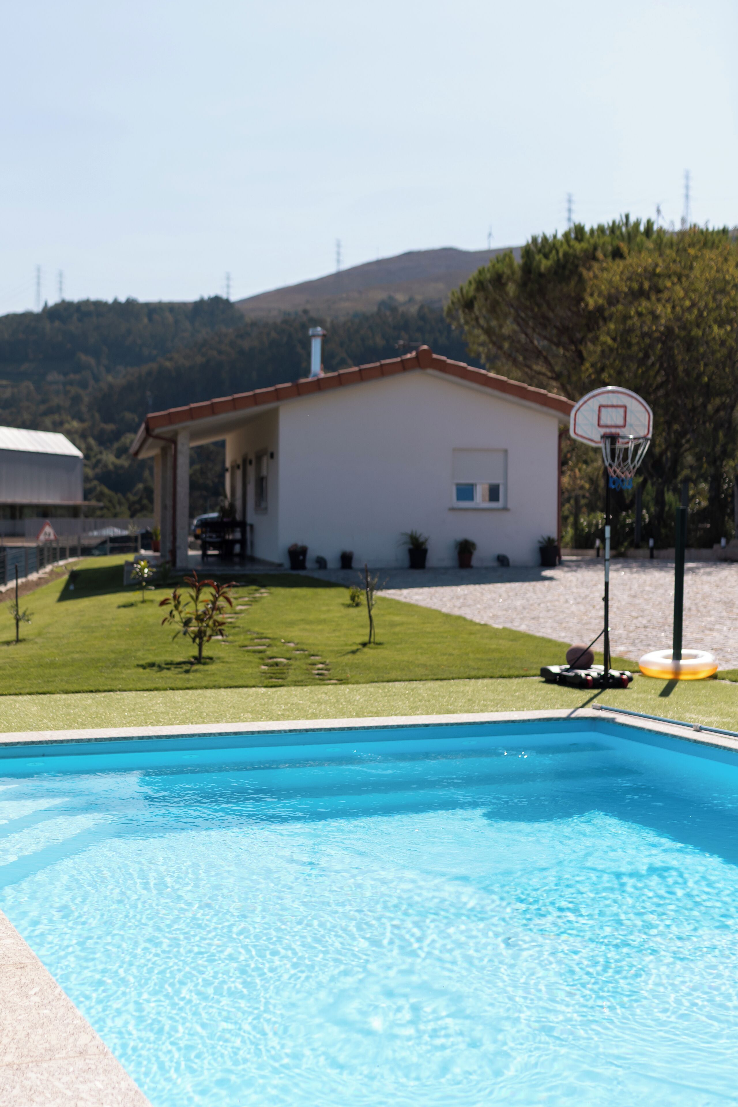 A swimming pool with a basketball hoop, a house, and a hilly landscape in the background.