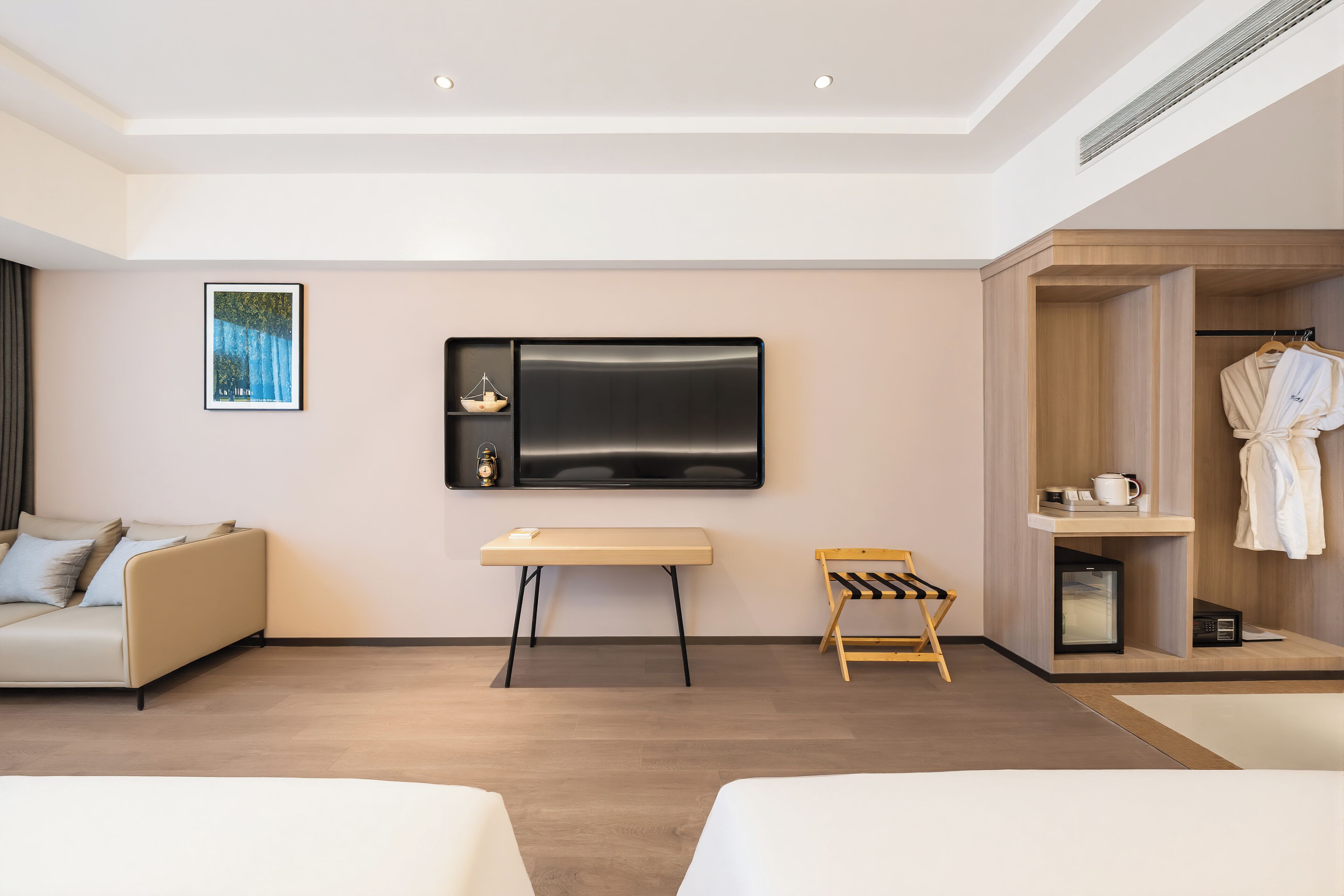 A modern hotel room with a sofa, a small table, a TV, a mini-fridge, a robe hanging, and a wooden bench.
