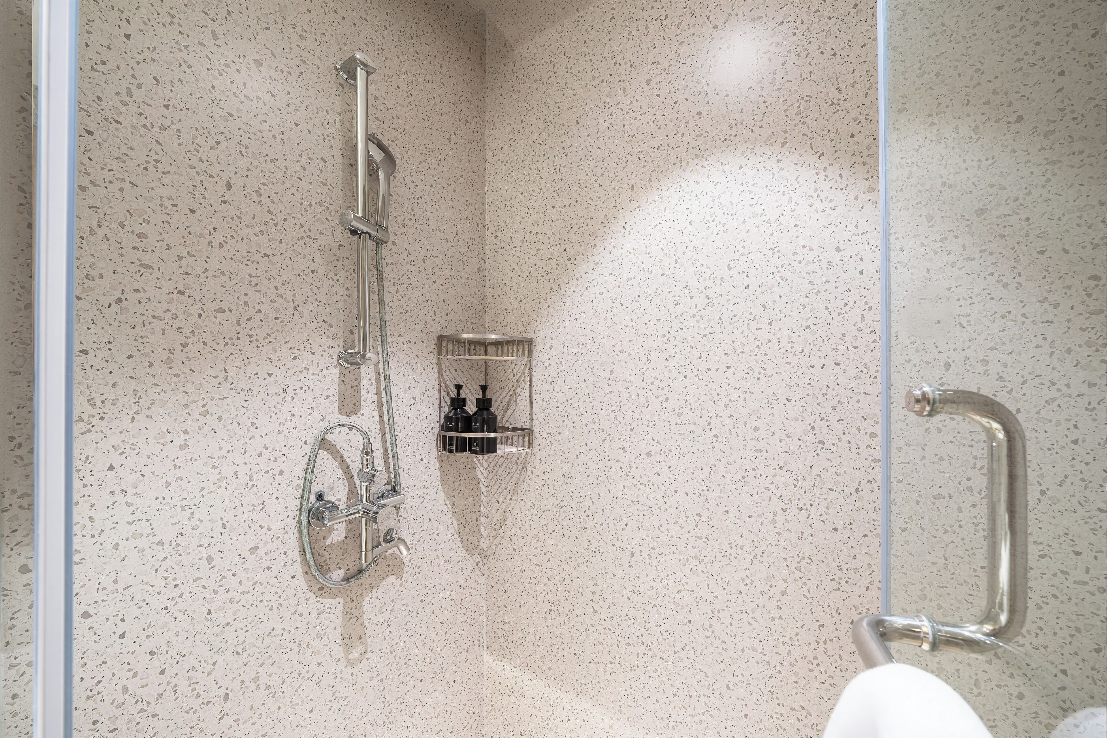 A shower area with a glass door, a wall-mounted handrail, and a shelf with toiletries.