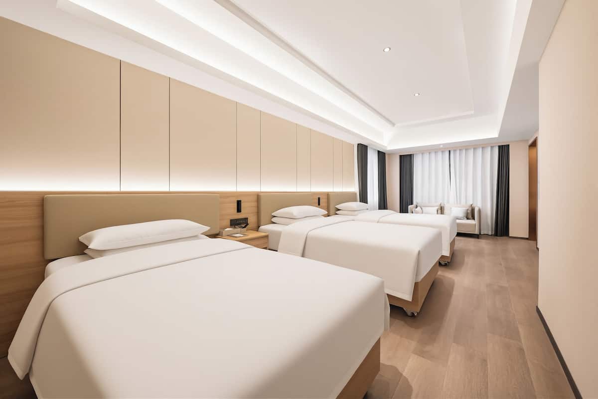 Quarto de hotel moderno com três camas, cabeceira de madeira, mesa de cabeceira e uma janela grande com cortinas.