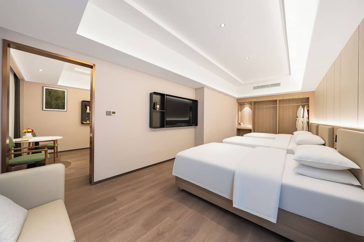 Quarto de hotel moderno com uma cama grande, televisão fixada na parede, guarda-roupa de madeira e uma pequena área de refeições com mesa e cadeiras.