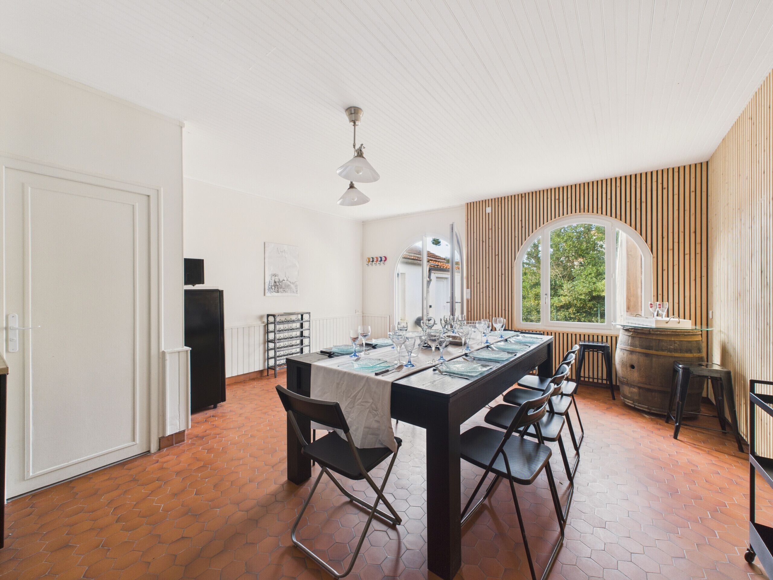 Een eetruimte met een lange tafel voor zes personen, omringd door stoelen, een houten vat en een groot raam met uitzicht op groen.