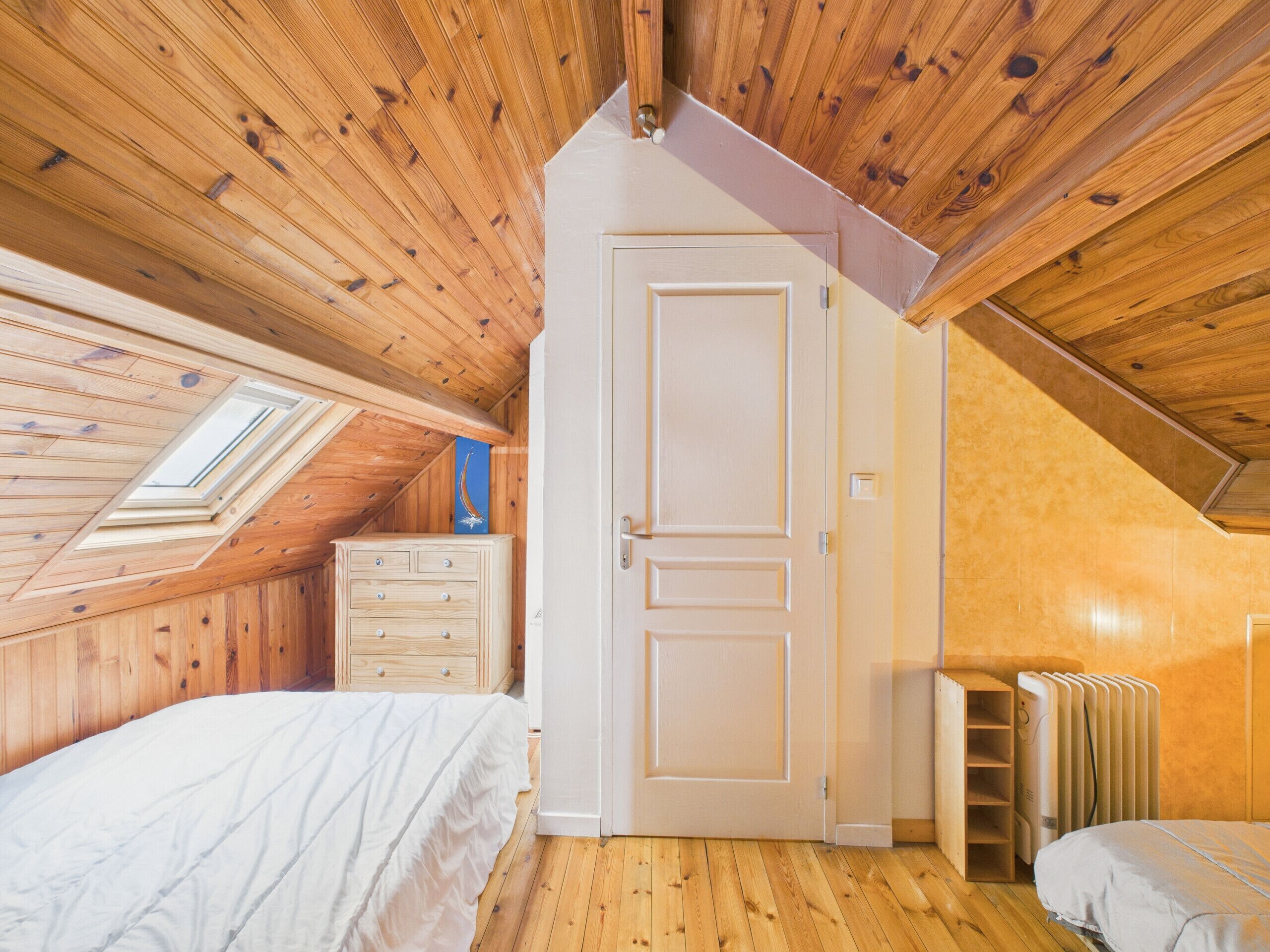 Une chambre avec des murs en bois, un plafond en pente, une porte blanche, une commode, un radiateur et un puits de lumière.