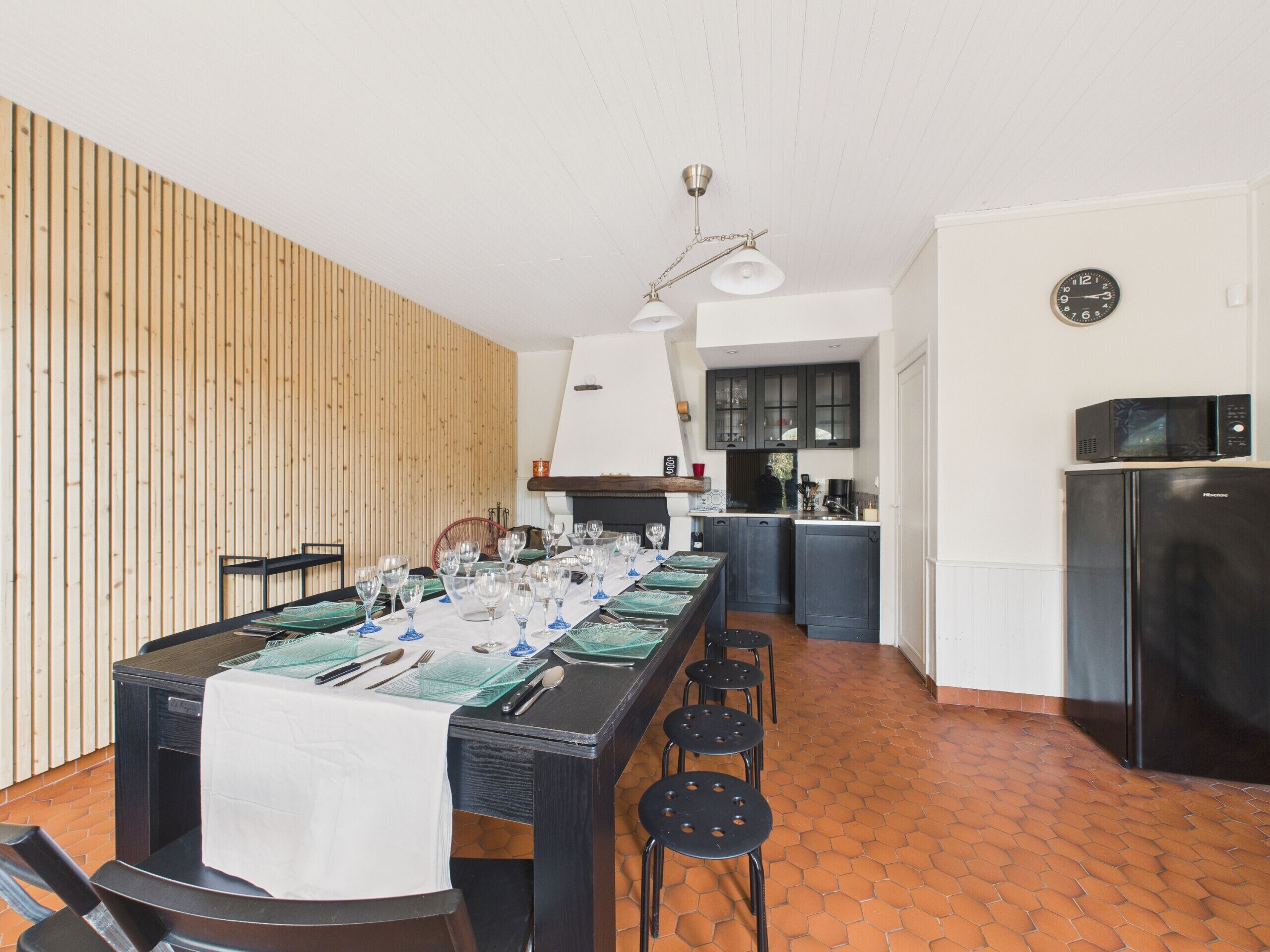 A dining area with a long table set for a meal, a kitchen in the background, and a clock on the wall.