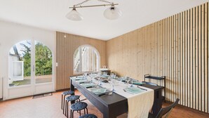 Een eetgedeelte met een lange tafel gedekt voor zes personen, omringd door zes krukken. De ruimte heeft houten lambrisering en een groot, rondbogig raam.