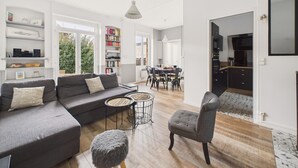Een moderne woonkamer met een grijze bank, een klein rond tafeltje en een grijze fauteuil. Er is een boekenplank vol boeken en een eethoek met een tafel en stoelen.