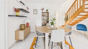 Un área de comedor moderna con una mesa de madera, sillas y una alfombra. Se ven estantes con objetos decorativos y una escalera al fondo.
