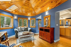 A room with a wooden ceiling, a sofa, a coffee table, a wooden cabinet, and a kitchen area with a sink and cabinets.